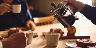 Artisan coffee being poured from a cafetiere into a mug on a table as friends sit around a table to chat and drink coffee.